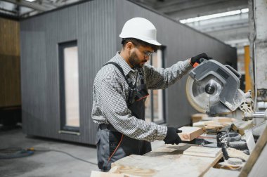 Modüler ev yapımı için testereyi ustalıkla kullanan bir inşaat atölyesinde çalışan işçiler, işçiliği ve hassasiyeti vurguluyor.