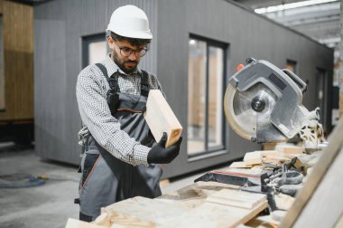 Marangoz prefabrik bir ev inşaatı atölyesinde, dairesel testerenin yanında ahşap bir kalas tutuyor ve kesmeye hazırlanıyor.