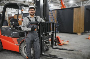 Arap mühendis, bir modüler ev üretim atölyesinde forklift yakınında dururken baş parmak hareketi yapıyor