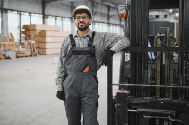 Güvenlik kaskı ve eldiven takan Arap depo işçisi modüler ev yapımı atölyesinde forklifte yaslanıyor.