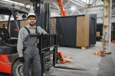 Arap forklift operatörü modüler ev yapım atölyesinde forkliftine yaslanmış pozlar veriyor.