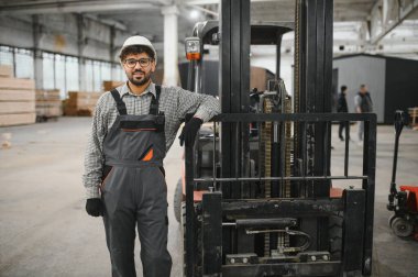 Modüler ev yapım atölyesinde forklifte yaslanmış, kameraya gülümseyen kendine güvenen Arap depo işçisinin portresi.