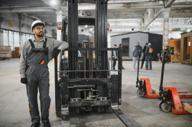 Arap inşaat işçisi. Tulumlu ve kasklı. Modüler bir ev fabrikasında forklifte yaslanıp üretimi denetliyor.