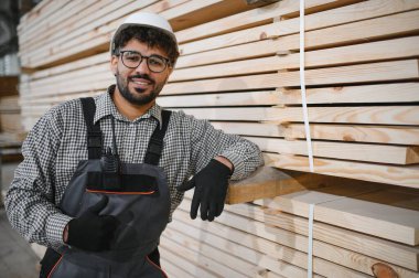 Kendine güveni tam bir Arap marangoz. İş kıyafeti ve başlık takıyor. Bir marangoz marangoz marangozluk fabrikasında kereste yığınına yaslanırken baş parmak hareketi yapıyor.