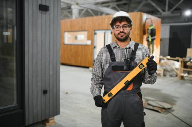 Kasketli ve tulumlu yetenekli inşaat işçisi bir ruh seviyesine sahip, bir fabrikada modüler ev üretimi sırasında kalite kontrol sağlıyor.