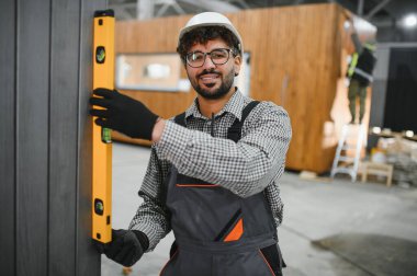 Gülümseyen Arap inşaatçı prefabrik ev atölyesindeki ruh seviyesiyle modüler ev duvarının dikey hizasını kontrol ediyor