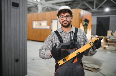 Güvenlik kaskı ve eldiven takan Arap inşaat işçisi ruh seviyesini koruyor ve bir modüler ev yapım atölyesinde durarak el kol hareketi yapıyor.