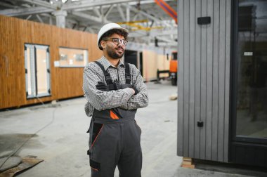 Kasketli ve tulumlu kendine güvenen inşaat işçisi, modüler bir ev fabrikasında çapraz silahlarla gururla duruyor, prefabrik inşaatlarda uzmanlık ve profesyonelliği somutlaştırıyor.