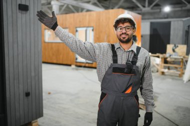 Güvenlik kaskı ve eldiven takan Arap inşaat işçisi, bir üretim atölyesinin içinde el kol hareketi yapıyor ve modüler bir ev gösteriyor.