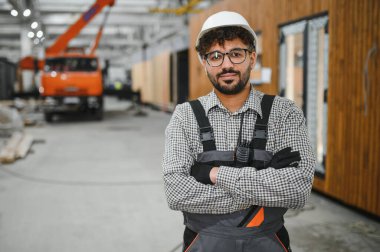 Kasklı ve eldivenli Arap inşaat işçisi prefabrik ev yapımı atölyesinde çapraz kollarla gülümsüyor.