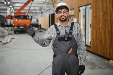 Güvenlik kaskı ve eldiven takan Arap inşaat işçisi modüler ev yapımı atölyesinde el kol hareketi yapıyor.