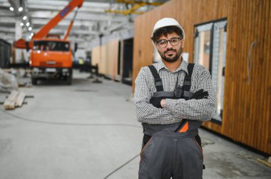 Prefabrik ev fabrikasında çapraz kollarla poz veren kendinden emin inşaat işçisi, modüler yapı yapımında uzmanlık gösteriyor.