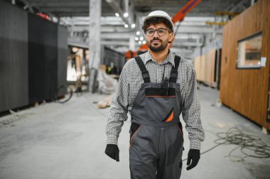 Arap inşaat işçisi modüler ev yapım atölyesinde yürüyor, güvenlik kaskı takıyor ve iş tulumu giyiyor.