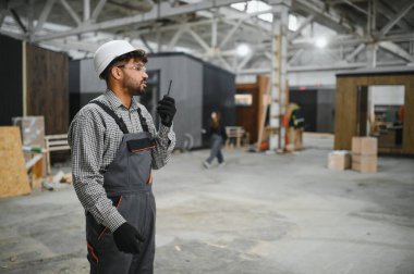 Arap inşaat işçileri prefabrik ev fabrikasında güvenlik kaskı takıyor ve telsiz kullanıyor.