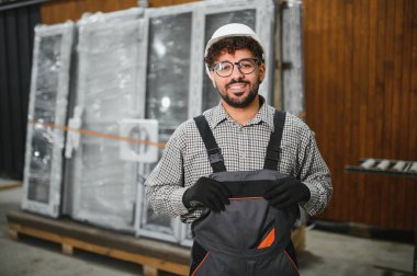 Güvenle gülümseyen, kasklı ve tulumlu bir Arap inşaat işçisi, modüler bir ev yapım atölyesinde uzmanlığını gösteriyor