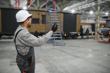 İnşaat işçisi, büyük bir endüstriyel atölyede modüler ev üretimini yönetirken telsiz kullanıyor ve el kol hareketi yapıyor.