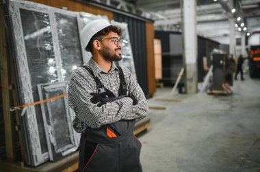 Güvenlik kaskı takan ve iş tulumu giyen genç bir inşaat işçisi prefabrik ev yapımı atölyesinde çapraz kollarla gülümsüyor.