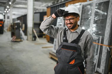 Arap inşaat işçisi, modüler ev yapım atölyesinde şapkasını düzeltirken gülümsüyor, işyerinden memnuniyet ve güvenlik gösterisi sergiliyor