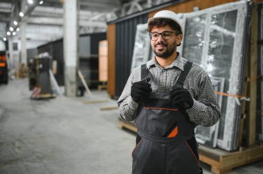 Güvenlik kaskı takan ve iş tulumu giyen Arap inşaat işçisi modüler ev yapım atölyesinde gülümsüyor