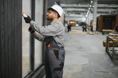 Modern bir fabrikada prefabrik modüler ev yapımı sırasında pencere çerçevesine dolgu macunu süren yetenekli bir işçi.