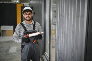 Arap inşaat işçisi silikon silah tutuyor, prefabrik ev atölyesinde modüler ev inşa ediyor.