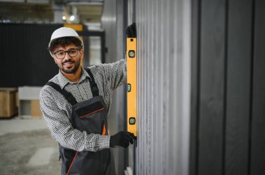 Arap inşaat işçisi, modüler ev fabrikasındaki prefabrik duvar panelinin dikey hizalanmasını sağlamak için, modern inşaattaki hassasiyet ve kalite kontrolünü vurgulayarak, ruh seviyesini kullanır.