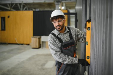 Arap inşaat işçisi, prefabrik inşaat atölyesinde modüler ev montajı sırasında duvar panelinin dikey hizalanmasını sağlayan bir ruh seviyesi kullanıyor.