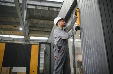 İnşaat işçisi prefabrikasyon atölyesindeki ruh seviyesini kullanarak modüler ev duvarının dikey hizalanmasını kontrol ediyor