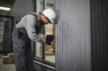 İnşaat atölyesinde prefabrik ev bölümünü monte eden elektrikli tornavida kullanan konsantre müteahhit.