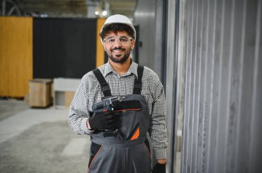Gülümseyen Arap inşaat işçisi güvenlik kaskı takıyor ve kablosuz matkap tutuyor, prefabrik modüler evi büyük bir atölyede monte ediyor.