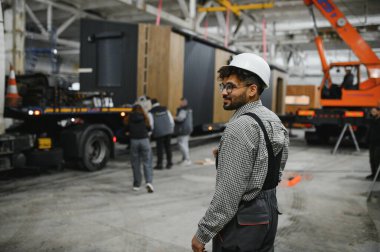 Sert bir şapka takan Arap mühendis, hareketli bir inşaat atölyesinde modüler evler üretimini denetliyor.