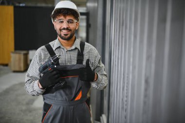 Güvenlik gözlüğü takan ve baret takan Arap inşaat işçisi, kablosuz matkap tutuyor ve modüler ev yapımı atölyesinde baş parmak hareketi yapıyor.