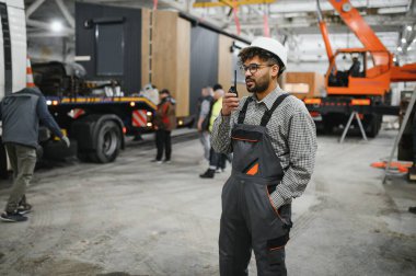 Güvenlik kaskı takan ve telsiz kullanan Arap mühendis modüler bir ev yapımı atölyesinde operasyonları koordine ediyor.