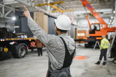 Güvenlik kaskı takan inşaat işçisi, modüler ev üretim fabrikasının içinde vinç operasyonlarını yönetirken parmağıyla işaret ediyor.