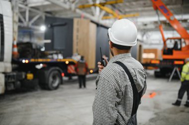 Arap mühendis telsiz kullanıyor, prefabrik modüler ev inşa atölyesinde operasyonları koordine ediyor.