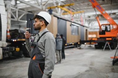 Güvenlik kaskı takan inşaat işçisi telsiz kullanıyor. Modüler bir ev üretim tesisinde operasyonları koordine ediyor.
