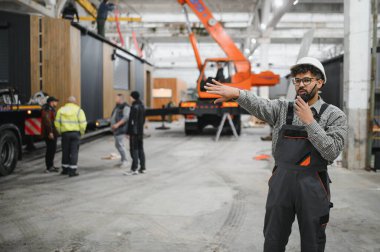 Güvenlik kaskı takan ve telsiz kullanan Arap mühendis yoğun bir inşaat atölyesinde modüler ev üretimini koordine ediyor.