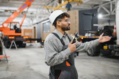 Güvenlik kaskı takan ve telsiz kullanan Arap mühendis, bir modüler ev inşaatı atölyesinde operasyonlar düzenliyor.