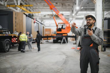 Arap mühendis telsiz kullanıyor ve inşaat atölyesinde prefabrik modüler ev yapımı sırasında işçilere talimat veriyor.