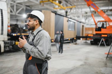 Güvenlik kaskı takan ve telsiz kullanan Arap mühendis, bir modüler ev inşaatı atölyesinde operasyonlar düzenliyor.
