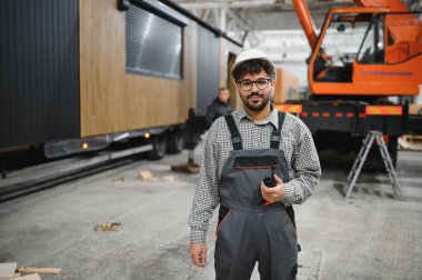 Arap mühendis elinde telsiz, bir inşaat atölyesinde prefabrik modüler ev üretimini denetliyor, verimli bir iş akışı ve kalite kontrolü sağlıyor.