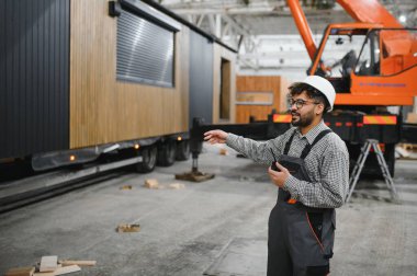 Arap inşaat işçisi sabit şapka takıyor ve tulum giyiyor. Modüler ev üretim tesisinde vinç kullanmak için telsiz kullanıyor.