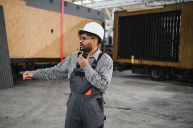 Güvenlik kaskı takan ve telsiz kullanan inşaat işçisi, modüler ev üretim tesisinde operasyonları koordine ediyor.