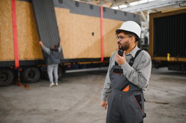 Arap mühendis telsiz kullanıyor, bir inşaat atölyesinde modüler ev ulaşımını koordine ediyor, prefabrik birimler kamyonlara yükleniyor.