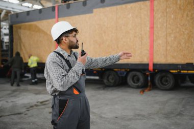 Arap mühendis telsiz kullanarak modüler ev ulaşımını koordine ediyor, bir inşaat atölyesinde talimatlar veriyor.