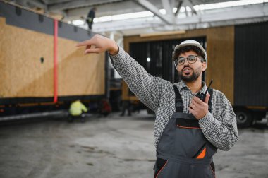 Arap inşaat işçisi, güvenlik kaskı takıyor ve telsiz kullanıyor. Yoğun bir atölyede modüler ev yapımını işaret ediyor ve yönetiyor.