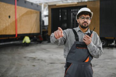 Üniforma giyen ve parmakla işaret eden Arap mühendis telsiz kullanarak prefabrikasyon atölyesinde modüler ev montajını koordine ediyor.