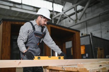 Odaklanmış Arap marangoz, bir marangoz fabrikasında modüler ev inşaatı sırasında hassasiyet ve kaliteyi sağlamak için ruh seviyesini kullanır.