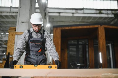 Odaklanmış marangoz ahşap seviyesini ruh seviyesiyle ölçüyor, parlak bir marangozluk atölyesinde prefabrik ev inşaatı üzerinde çalışıyor.