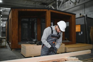 Arap inşaat işçisi elektrikli tornavida kullanıyor, büyük atölyede prefabrik modüler ev için ahşap parçalar birleştiriyor.